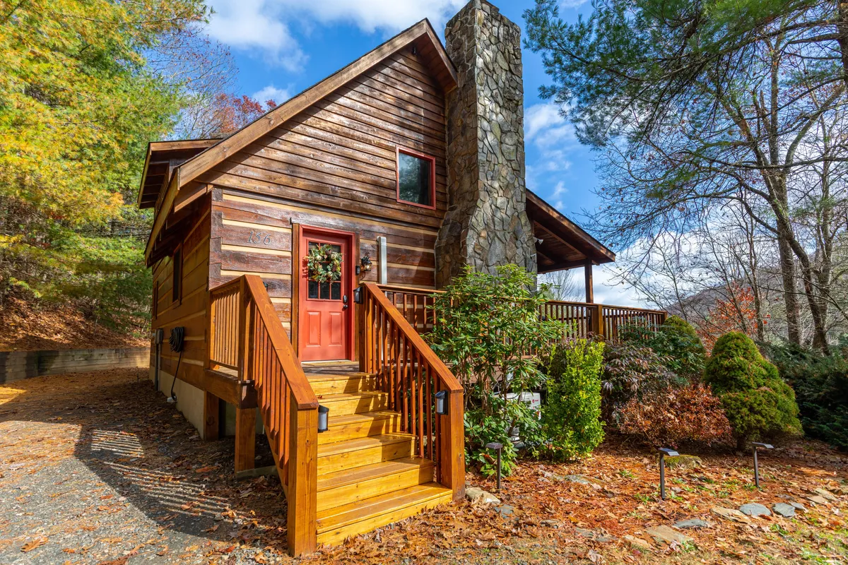 The image shows the inviting exterior of a cozy log cabin nestled among lush foliage, featuring a charming wooden front door, a rustic stone chimney, steps leading up to a welcoming porch, and hints of mountain views beyond. The scene exudes warmth and tranquility, perfect for relaxing on the porch or gathering with loved ones as the leaves drift by.