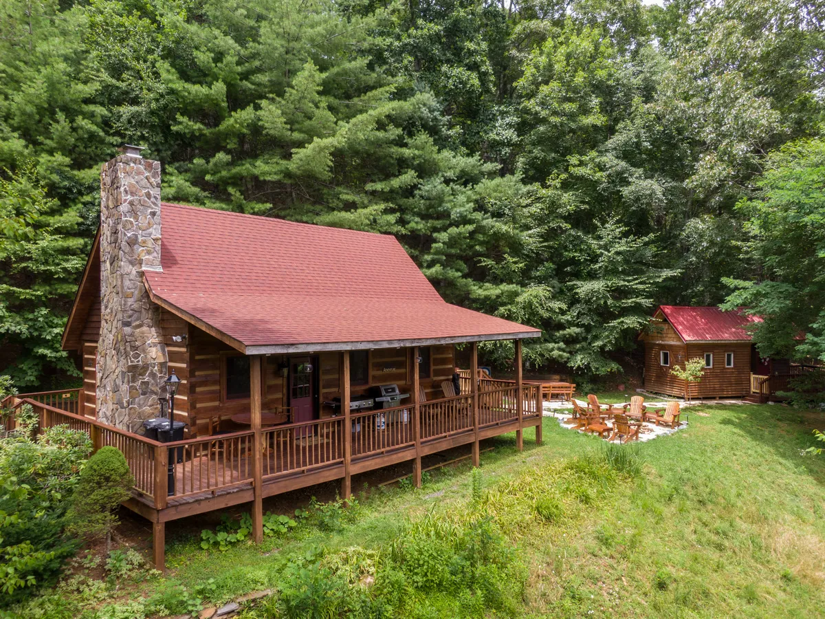 A cozy log cabin surrounded by tall trees, with a spacious deck and a stone chimney. The fire pit area is set up for gatherings, and the surrounding natural landscape hints at nearby trails and open spaces.