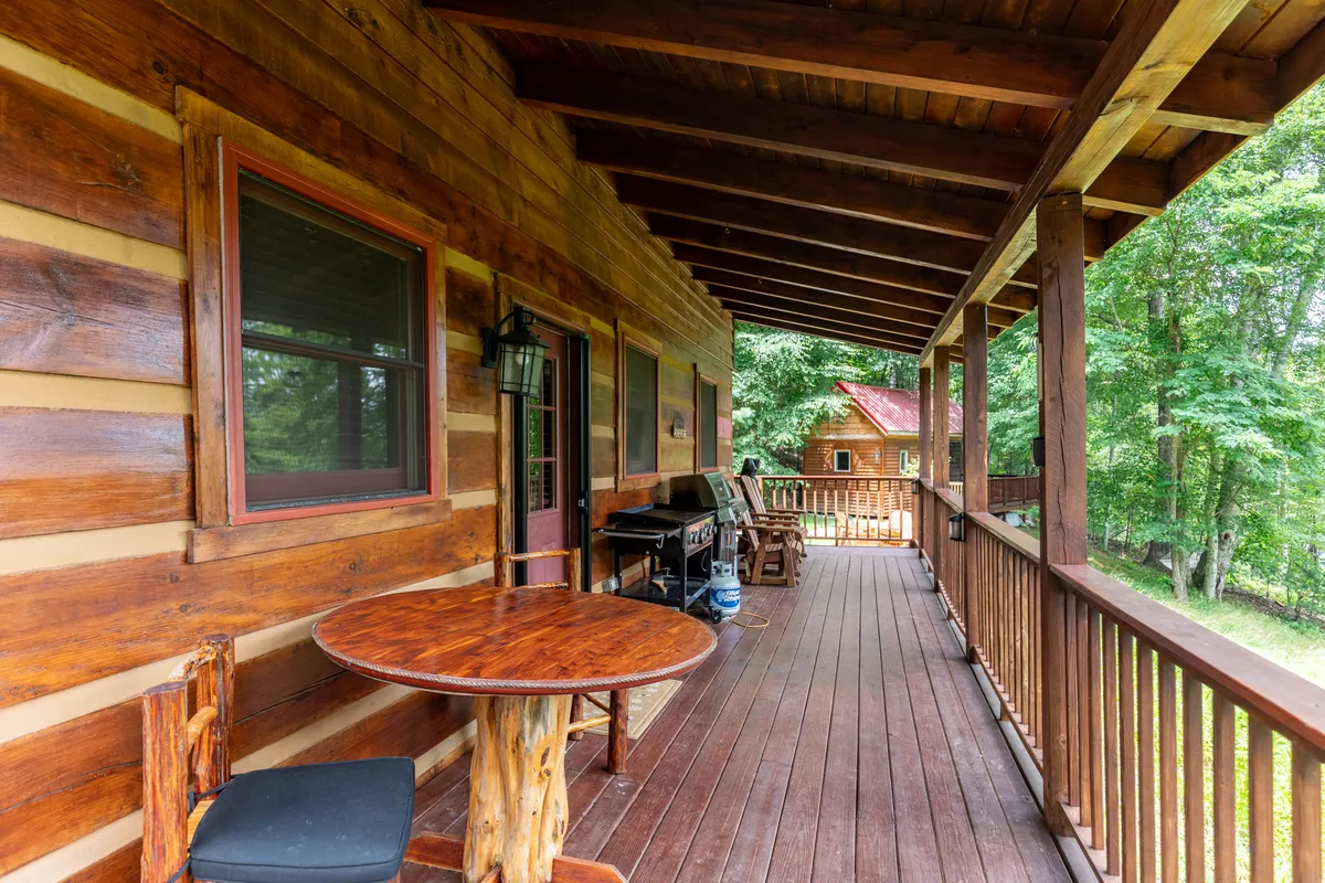 The image highlights a wooden deck with a table and chairs, a grill, and lush surrounding greenery. The atmosphere is cozy, with opportunities for outdoor dining, socializing, and enjoying fresh mountain air.