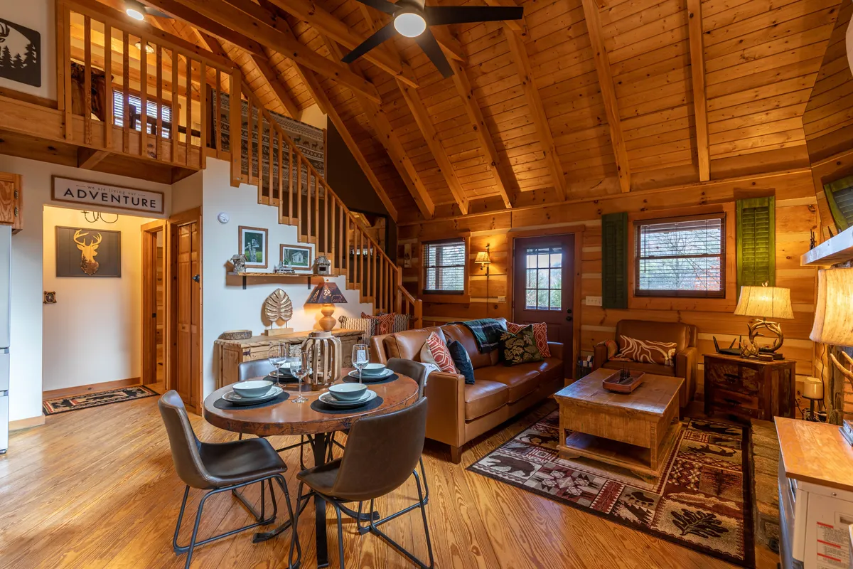 A warm interior scene displaying a round dining table set for a small gathering, a comfortable sofa, decorative wall art, and a classic wooden ceiling. The setting is intimate, perfect for both relaxing and connecting with family and friends.