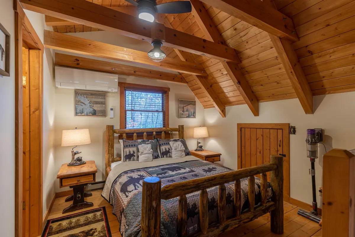 A beautiful log bed dominates this warmly lit bedroom. The space features bedside tables, decorative pillows, a wood-paneled ceiling, and a window that lets in natural light and frames peaceful views of the outdoors. The rustic charm and plush bedding make the room feel inviting and restful.