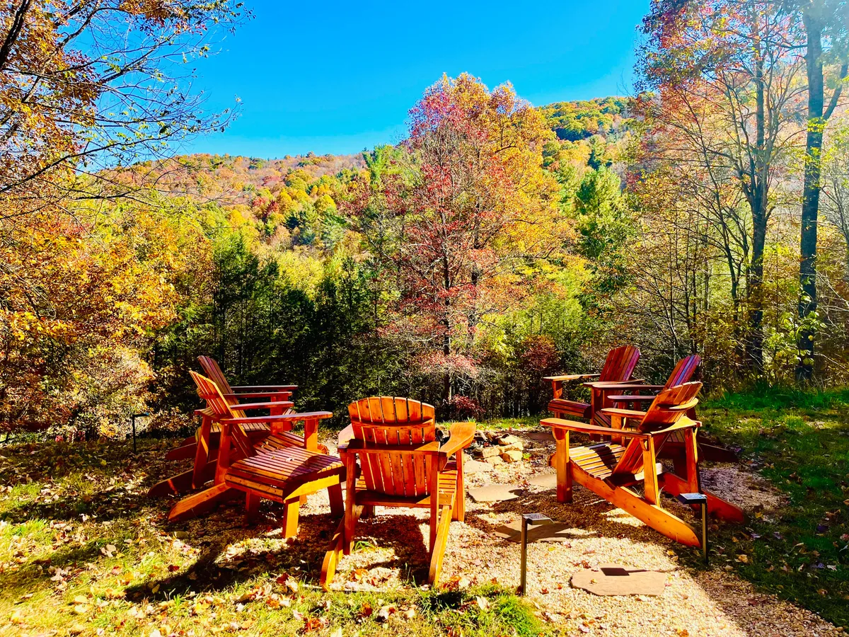 This exterior view features Adirondack chairs arranged around a fire pit, set amidst towering trees with a sweeping view of the mountains beyond. The scene is ideal for gathering, watching sunsets, or enjoying crisp evenings by the warm fire.