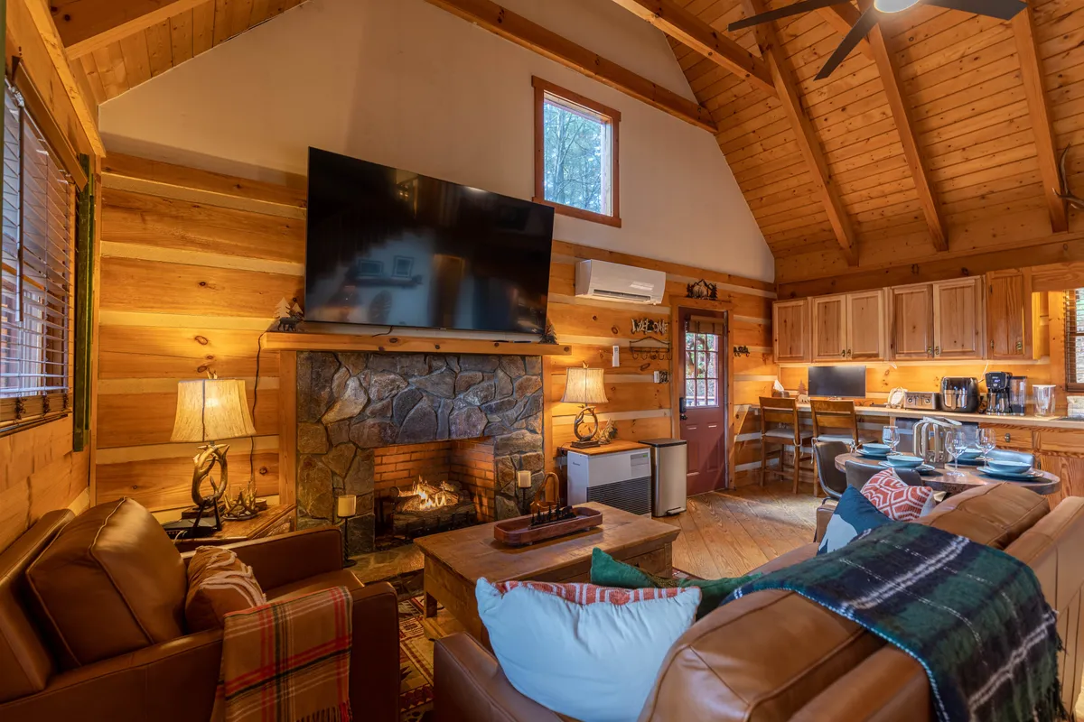 This image showcases the main living area of the cabin, featuring a striking stone gas fireplace, a large television, exposed wooden beams, leather sofas, and an open kitchen area in the background. The overall atmosphere is warm, inviting, and perfect for social gatherings.