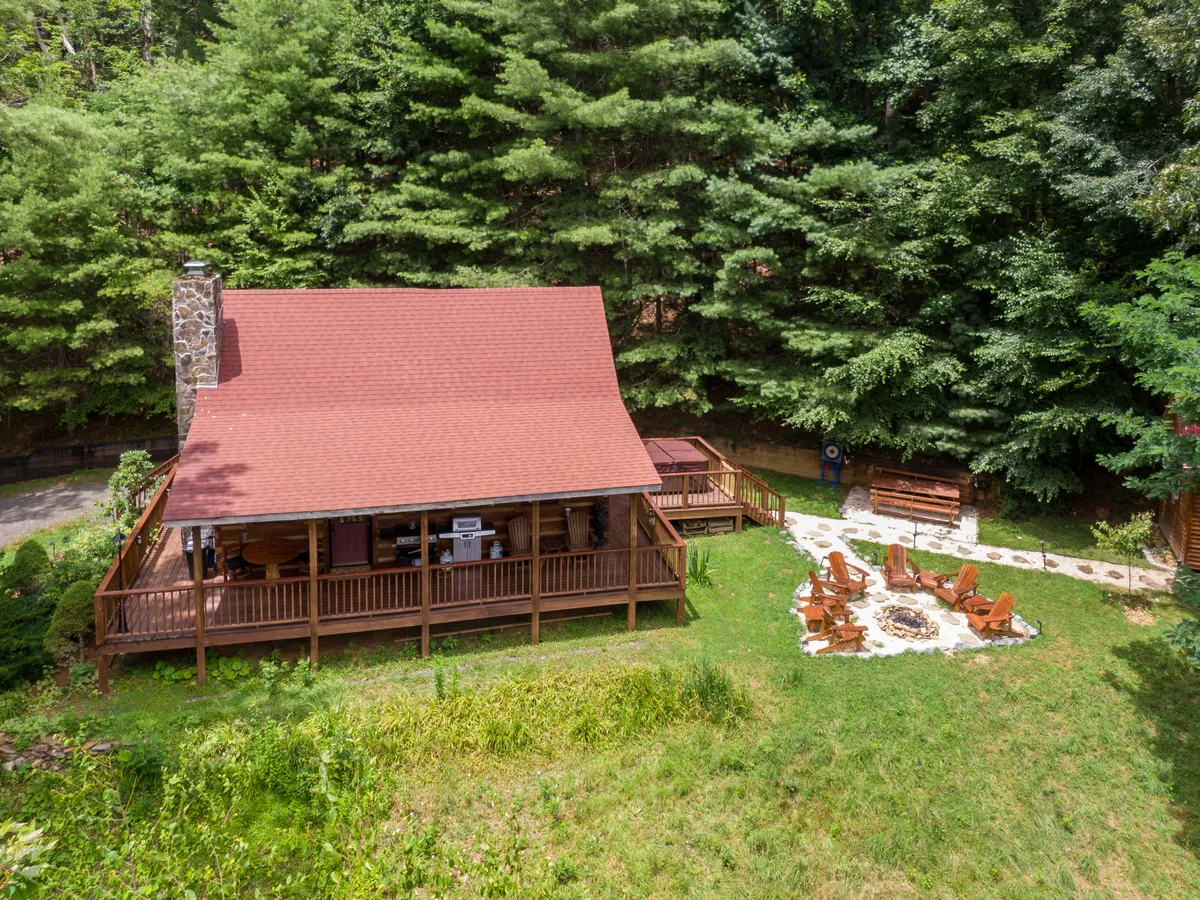 A rustic fire pit surrounded by six Adirondack chairs, set against a wooded backdrop next to a charming wooden cabin. The setup is ideal for campfire gatherings, storytelling, and marshmallow roasting under the stars.
