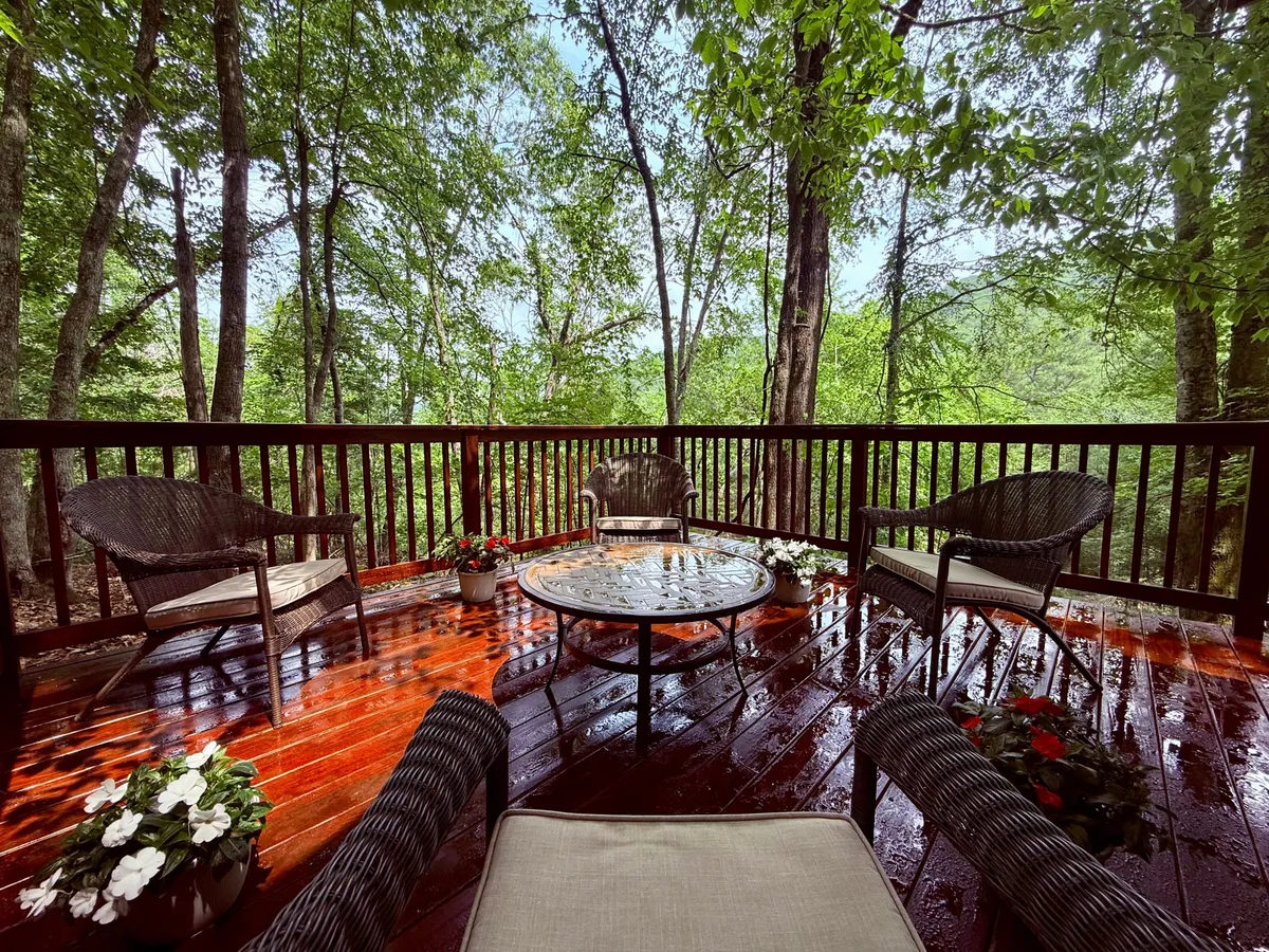 An inviting outdoor seating area with a glass table is surrounded by lush greenery and colorful flower pots, offering a peaceful spot to relax, read, or socialize. The vibrant surroundings hint at the beauty of spring, summer, and early fall in the mountains.