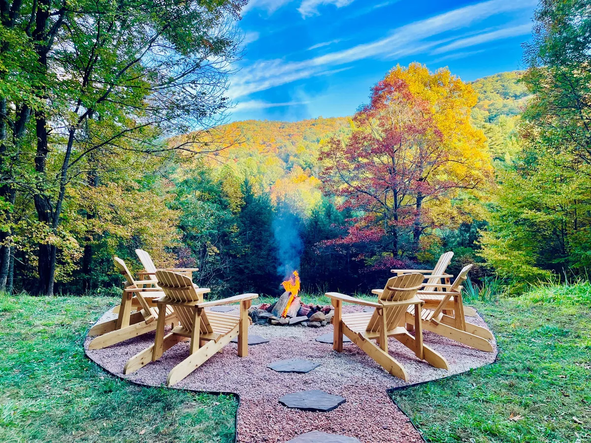 This exterior scene features a cozy fire pit surrounded by wooden chairs with a clear view of vibrant mountain scenery in the background. The gathering area is nestled among trees, offering a welcoming spot for relaxation after a day of exploring.