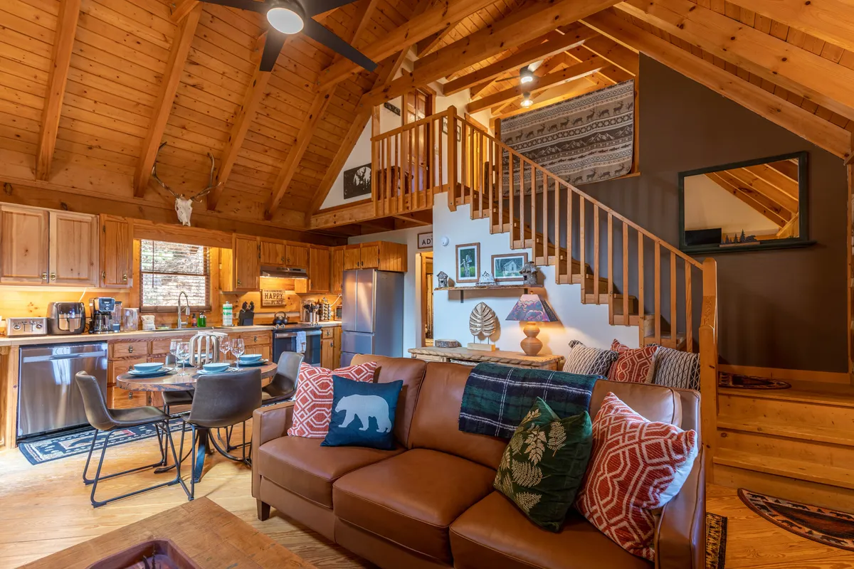 The image features a welcoming open-plan space with wooden beams overhead, a kitchen area, a dining table, and a cozy sofa with patterned cushions. The scene feels homey and ideal for group activities such as shared meals and game nights.