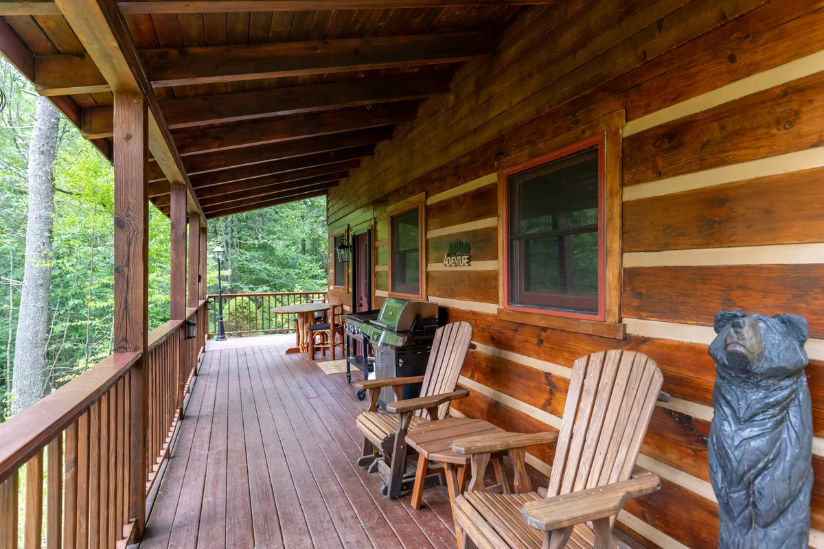 A covered porch featuring classic wooden rocking chairs, a barbecue grill, and a unique wooden bear sculpture. The porch looks out onto serene greenery, inviting guests to savor their morning coffee or unwind in the fresh air.