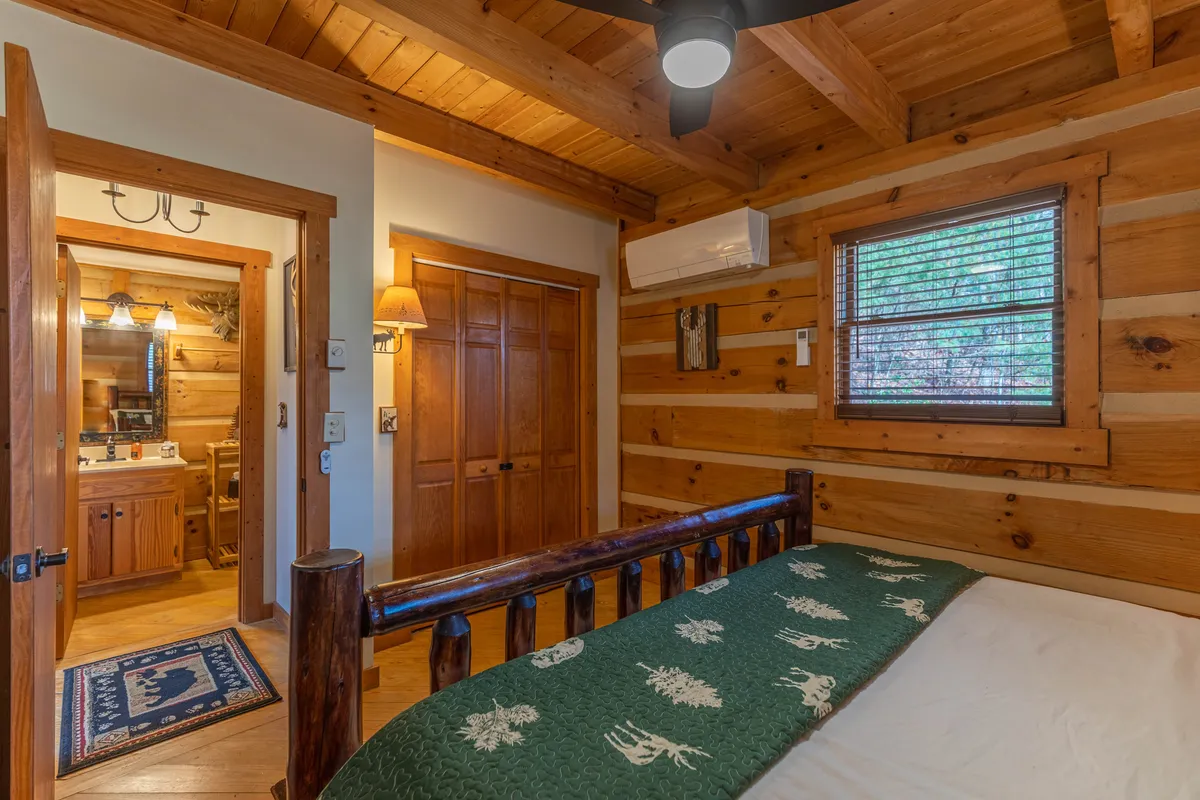 A wooden bed frame with a green patterned blanket anchors a cozy loft bedroom. Wood paneling and a window with soft, natural light set a tranquil scene, while a doorway leads to a bathroom, hinting at ensuite convenience. The space exudes warmth and peacefulness, ideal for a relaxing stay.