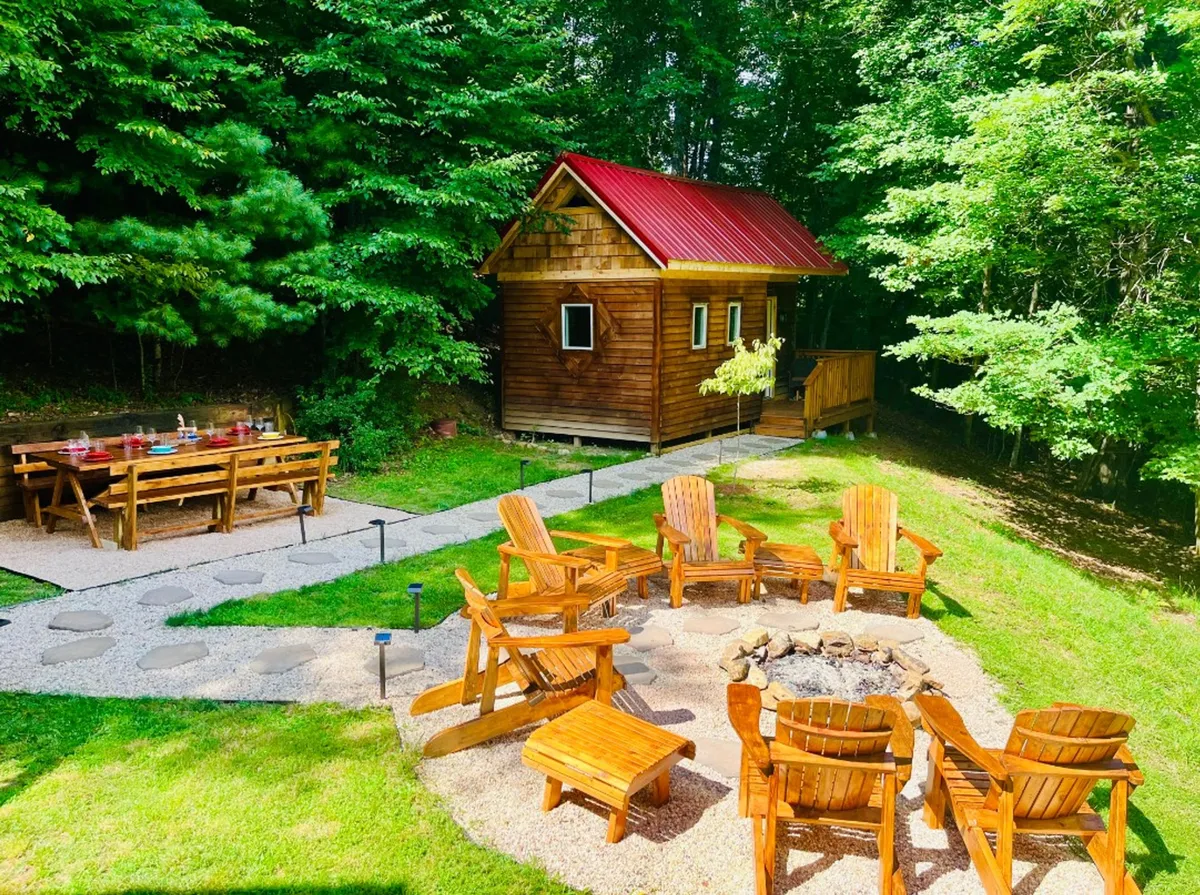 A cozy outdoor gathering area in front of a wooden cabin, with a fire pit encircled by Adirondack chairs, a dining table for shared meals, and the forest in the background. The space invites social gatherings, storytelling, and stargazing under the open sky.