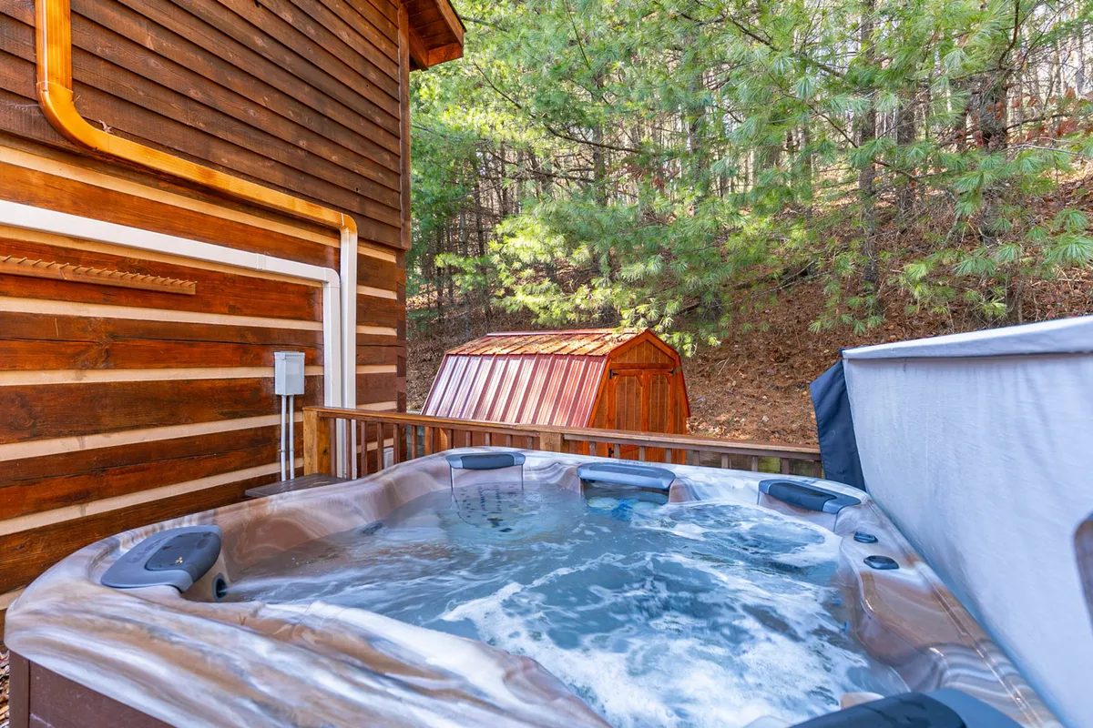 The image captures a tranquil exterior scene featuring a wooden cabin nestled among trees, a bubbling hot tub in the foreground, and a small shed nearby. The setting exudes relaxation with natural surroundings and the promise of enjoying crisp mountain air, whether during snowy winters or lush green summers.