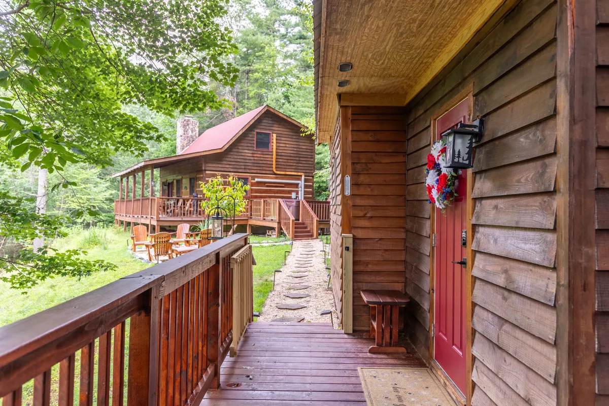 A welcoming wooden walkway lined with lush greenery leads to a red front door. A cozy seating area is visible, blending seamlessly with the natural setting and providing a quiet spot for both humans and dogs.